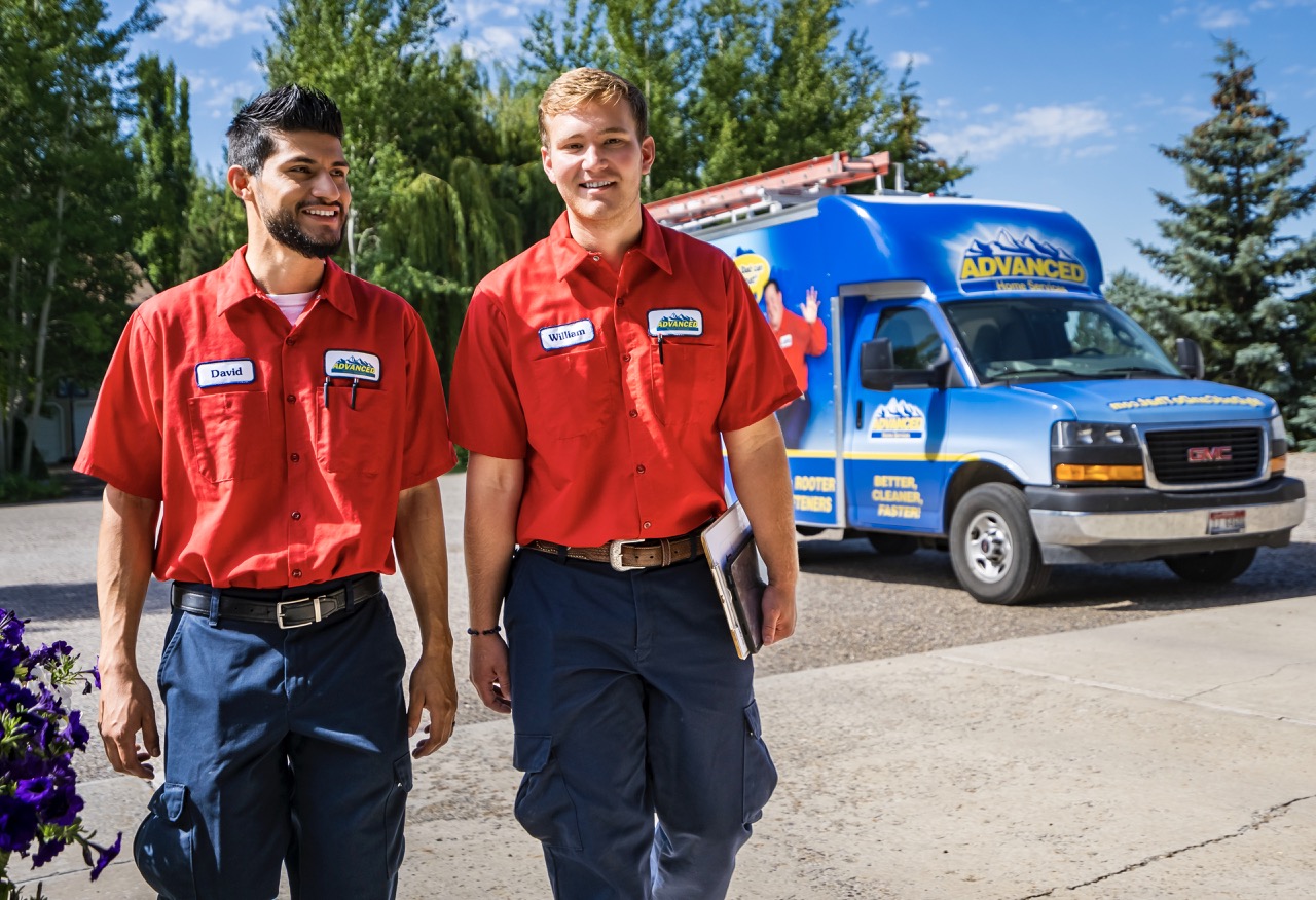Two Advanced Home Services technicians in red work shirts with name tags and navy blue work pants. Service truck and trees behind them.